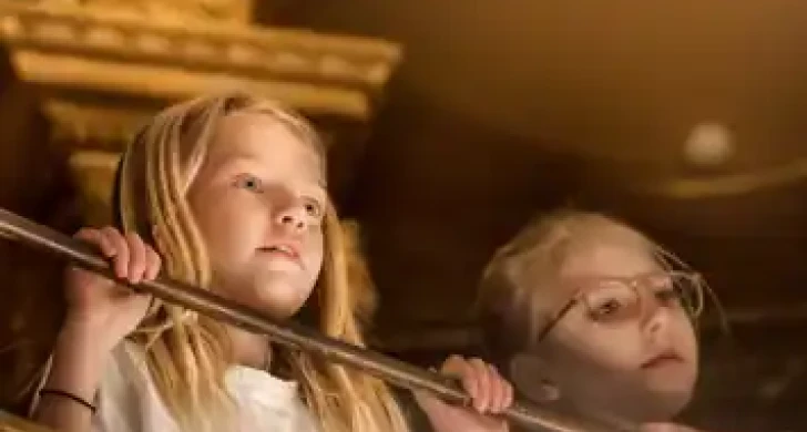 Två personer med suddade ansikten står bakom ett räcke inomhus, omgivna av varma toner.