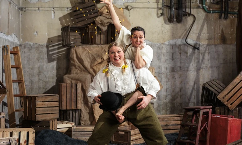 Två kvinnor, den ena sitter på den andras axlar och kastar konfetti, i ett källarrum fullt av trälador och annat bråte