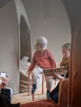 En kvinna läser en bok för ett barn som sitter på en pall. En annan kvinna sitter och tittar på sin mobil.