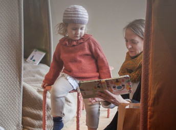 Ett barn sitter på en pall och en kvinna sitter strax nedanför och läser en bok. I bakgrunden och förgrunden är det textila hus.