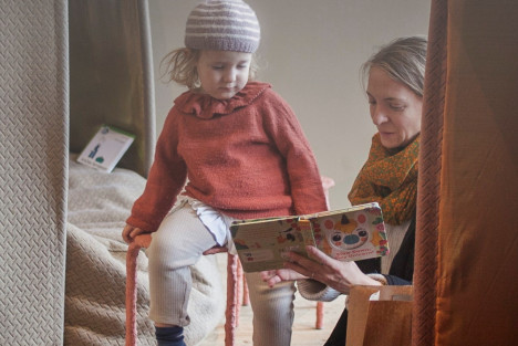 Ett barn sitter på en pall och en kvinna sitter strax nedanför och läser en bok. I bakgrunden och förgrunden är det textila hus.