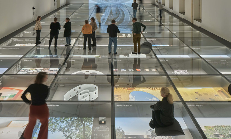 Bild på en stor sal med glasgolv. Under glasgolvet är det bilder och modeller. Människor står och sitter på glaset
