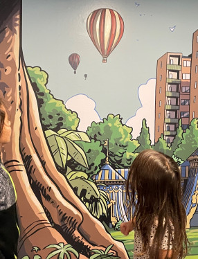 En pojke nio år och en flicka fem år tittar på en vägg med teckningar av en luftballong som flyger över ett fantasilandskap.