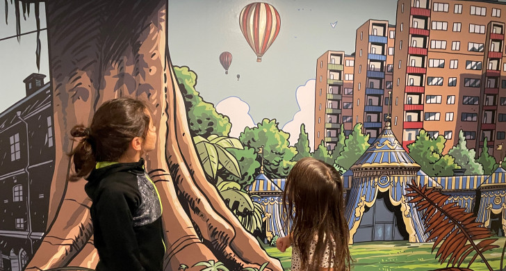 En pojke nio år och en flicka fem år tittar på en vägg med teckningar av en luftballong som flyger över ett fantasilandskap.
