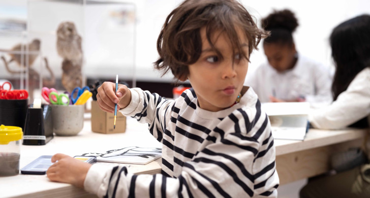 Ett barn som sitter vid ett bord och skapar. Han håller en penna i handen. och har precis vänt sig om.
