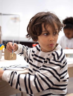 Ett barn som sitter vid ett bord och skapar. Han håller en penna i handen. och har precis vänt sig om.