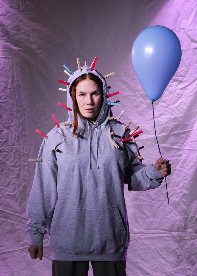Lily iklädd grå luvtröja håller i en blå ballong. På luvtröjan finns många klädnypor fastsatta.