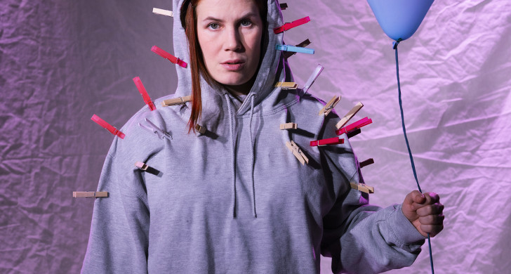 Lily iklädd grå luvtröja håller i en blå ballong. På luvtröjan finns många klädnypor fastsatta.