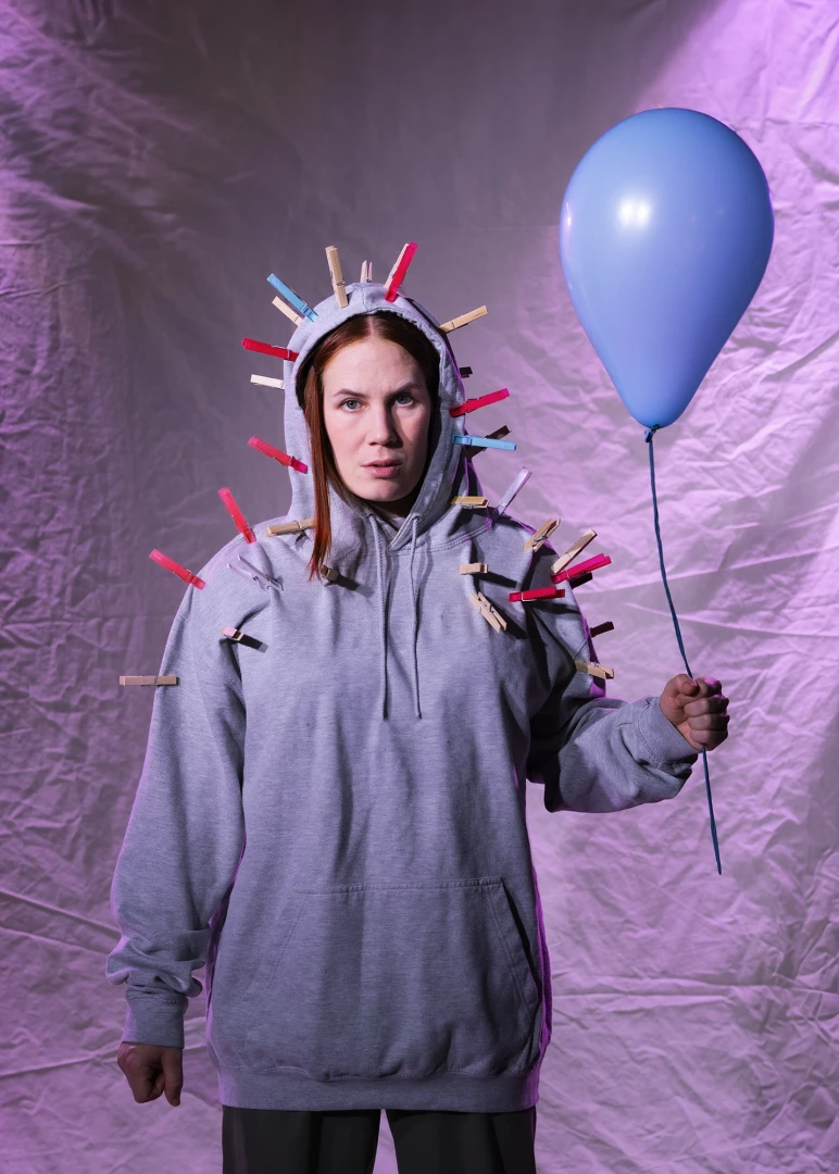 Lily iklädd grå luvtröja håller i en blå ballong. På luvtröjan finns många klädnypor fastsatta.