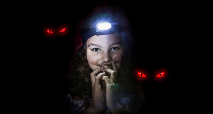 Flicka som tittar in i kameran med pannlampa på huvudet. Röda ögon syns i bakgrunden